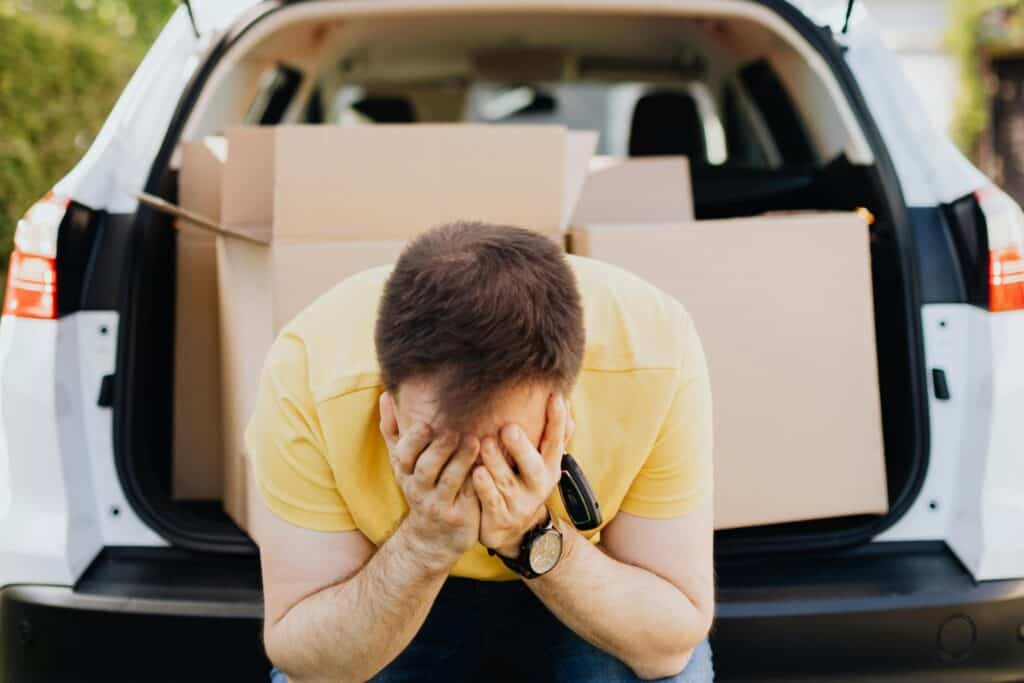 A man with his head on his palms sitting at the back of his car with cardboard