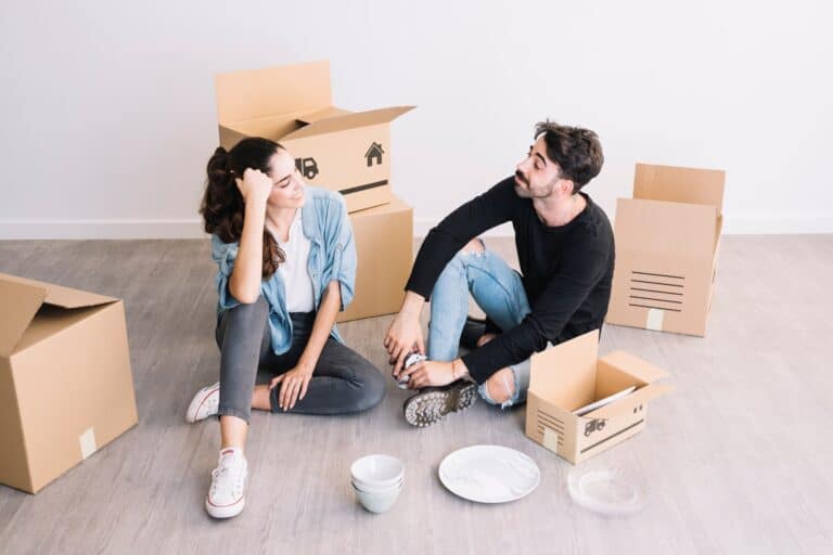 A young couple sitting on the floor of their new flat with opened boxes in the concept of 'what are the hidden costs in home removals services'.