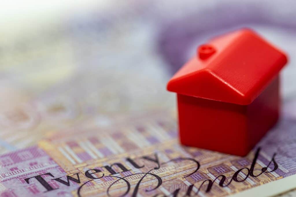 A red miniature house on top of a British pound note in the concept of temporary accommodation cost during the move - one of the hidden costs when moving